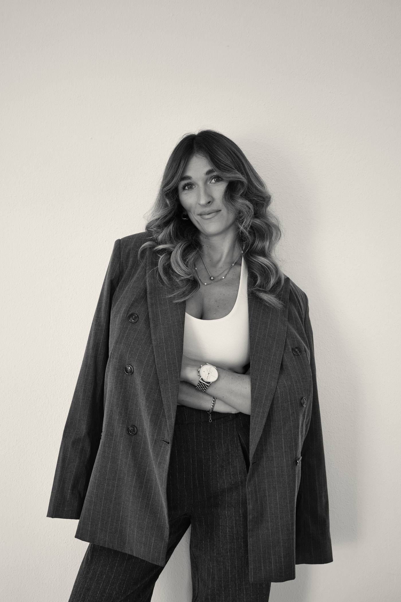 Steffi Cebis lehnt mit verschränkten Armen an einer hellen Wand, den Nadelstreifen-Blazer über die Schultern gelegt, und lächelt in die Kamera.